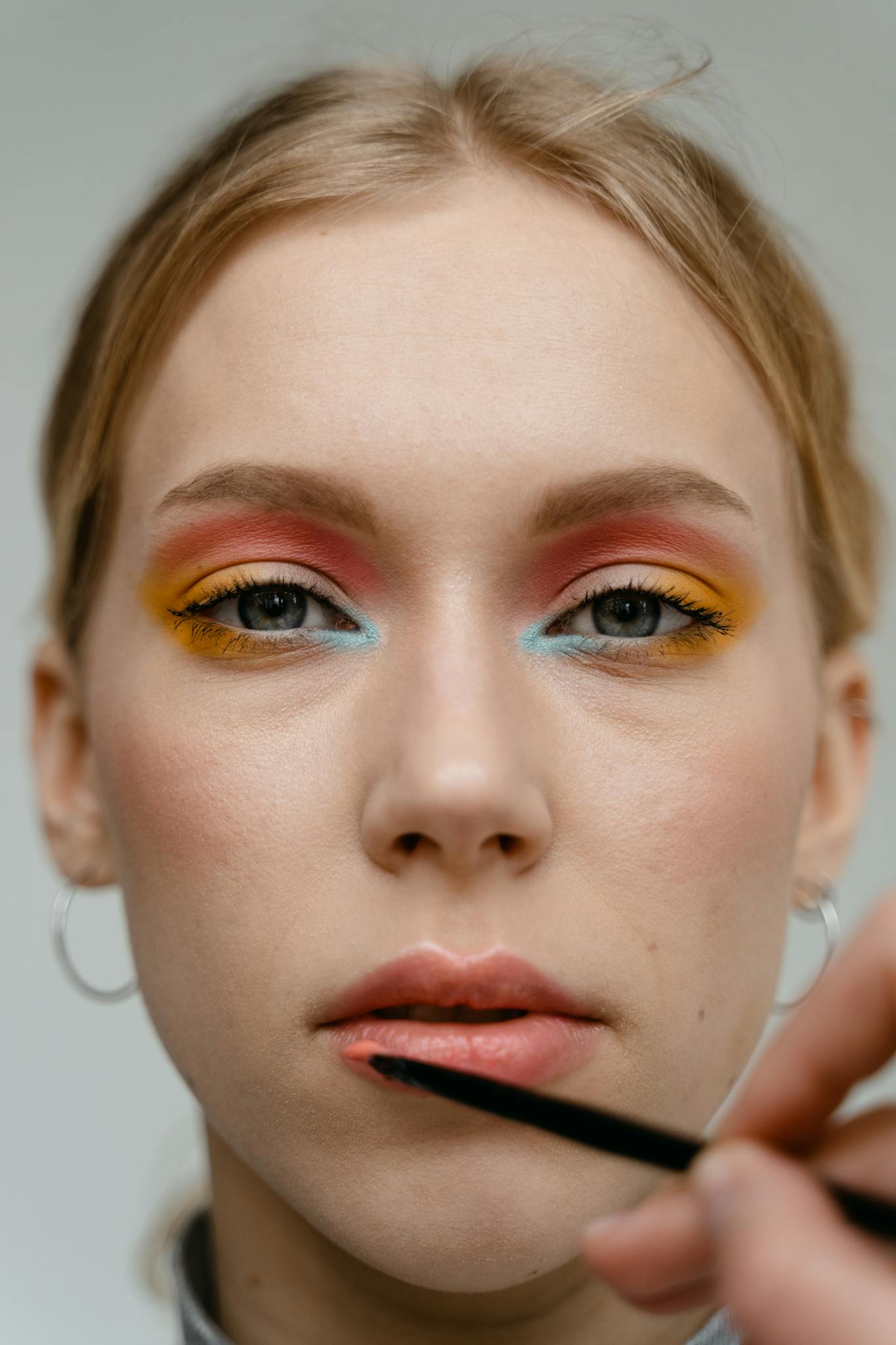 A woman with vibrant eyeshadow gets lipstick applied in a close-up shot.