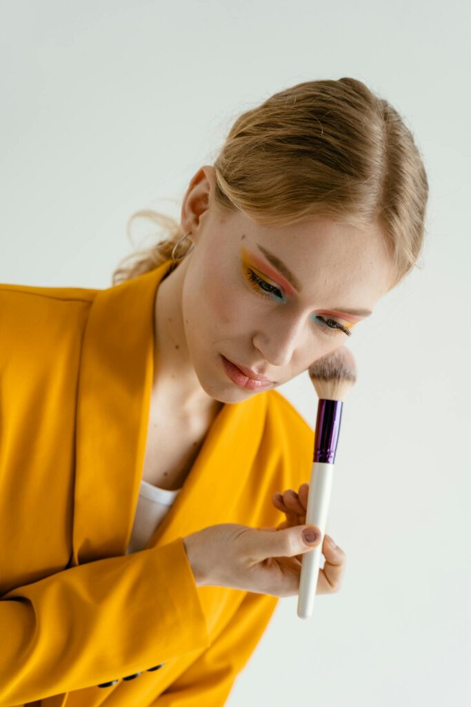 Close-up of a woman applying makeup with a brush, wearing bright yellow attire.